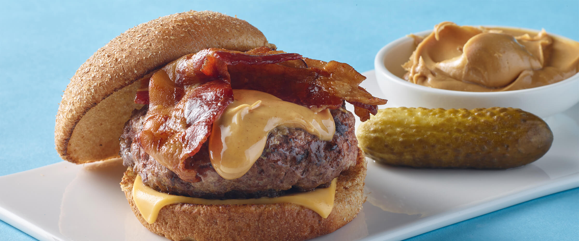 Bacon Peanut Butter Burger next to a pickle and a small dish of SKIPPY® peanut butter on a white plate