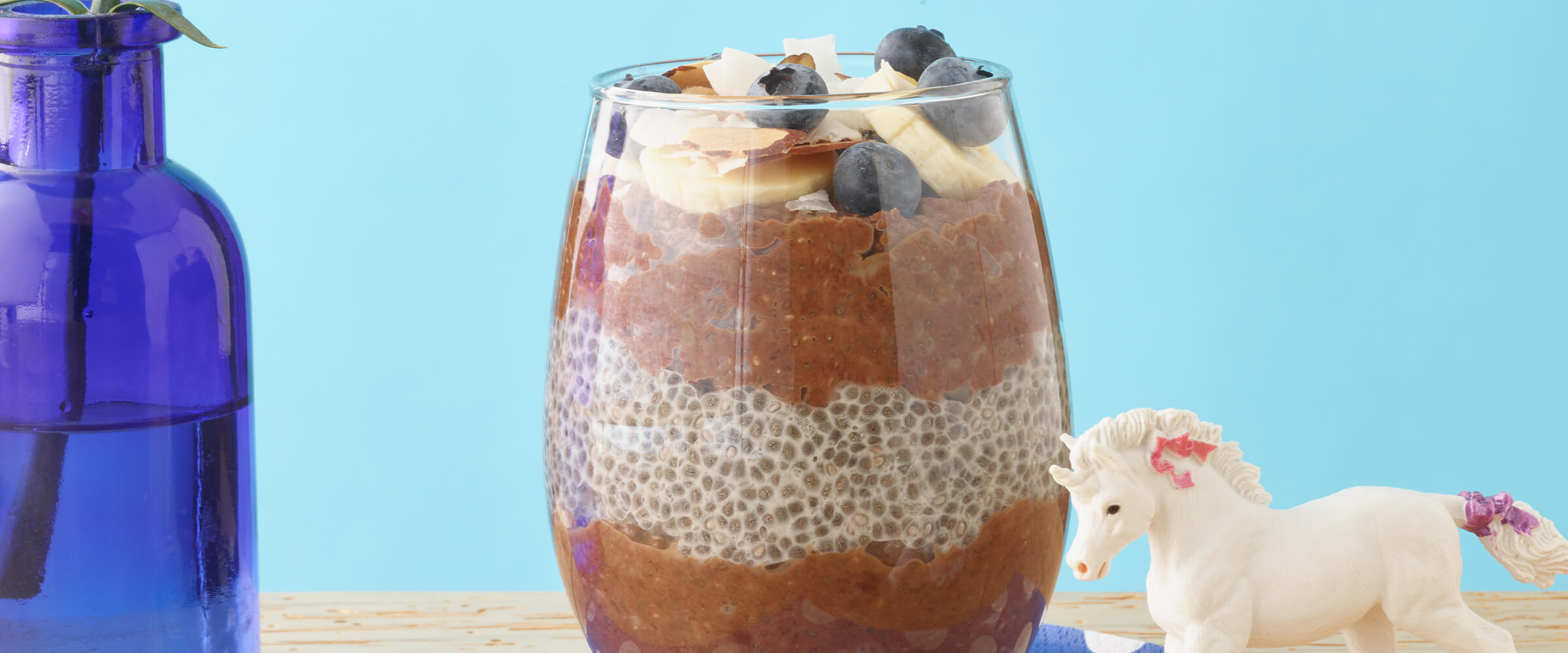 Peanut butter cocoa chia seed pudding parfaits on a wooden table with a unicorn figurine next to it on a teal background.