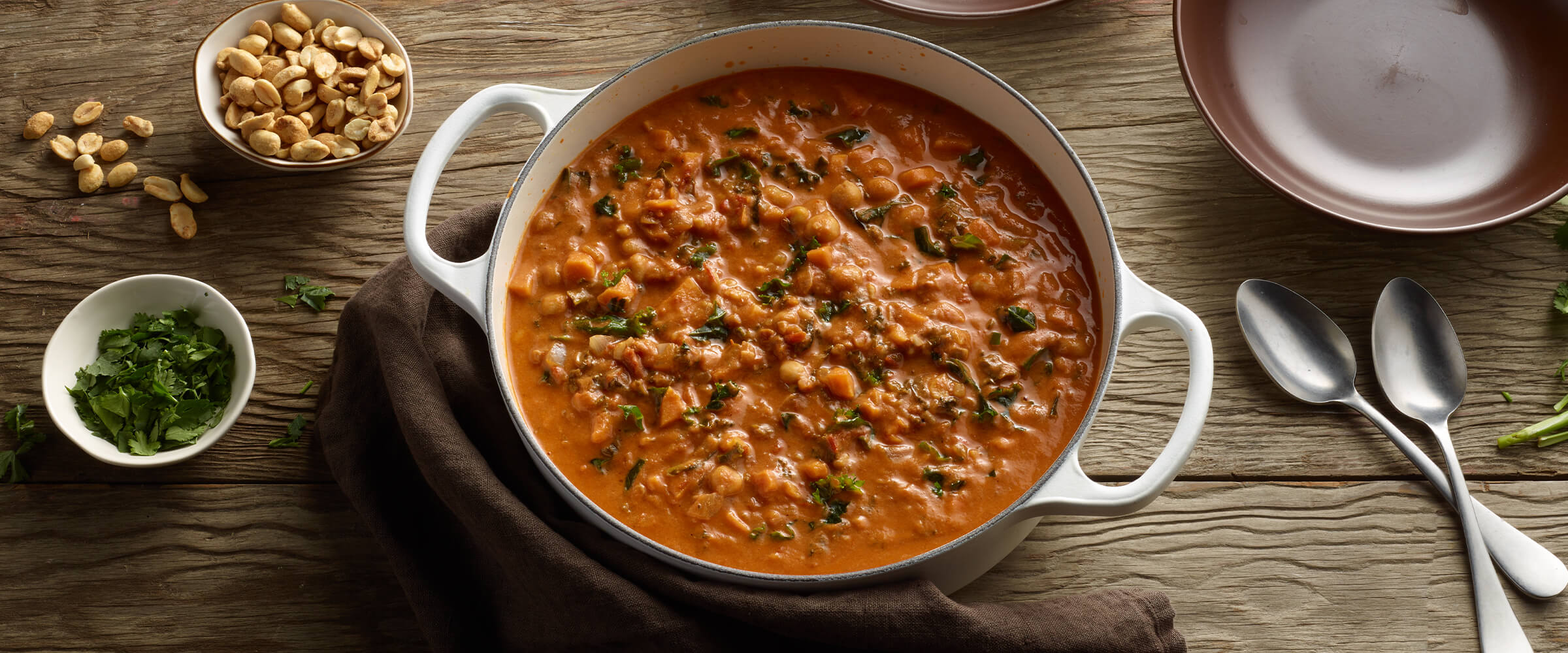 African peanut soup in a large pot with a small bowl of peanuts and herbs next to the pot. The pot is sitting on a wooden table