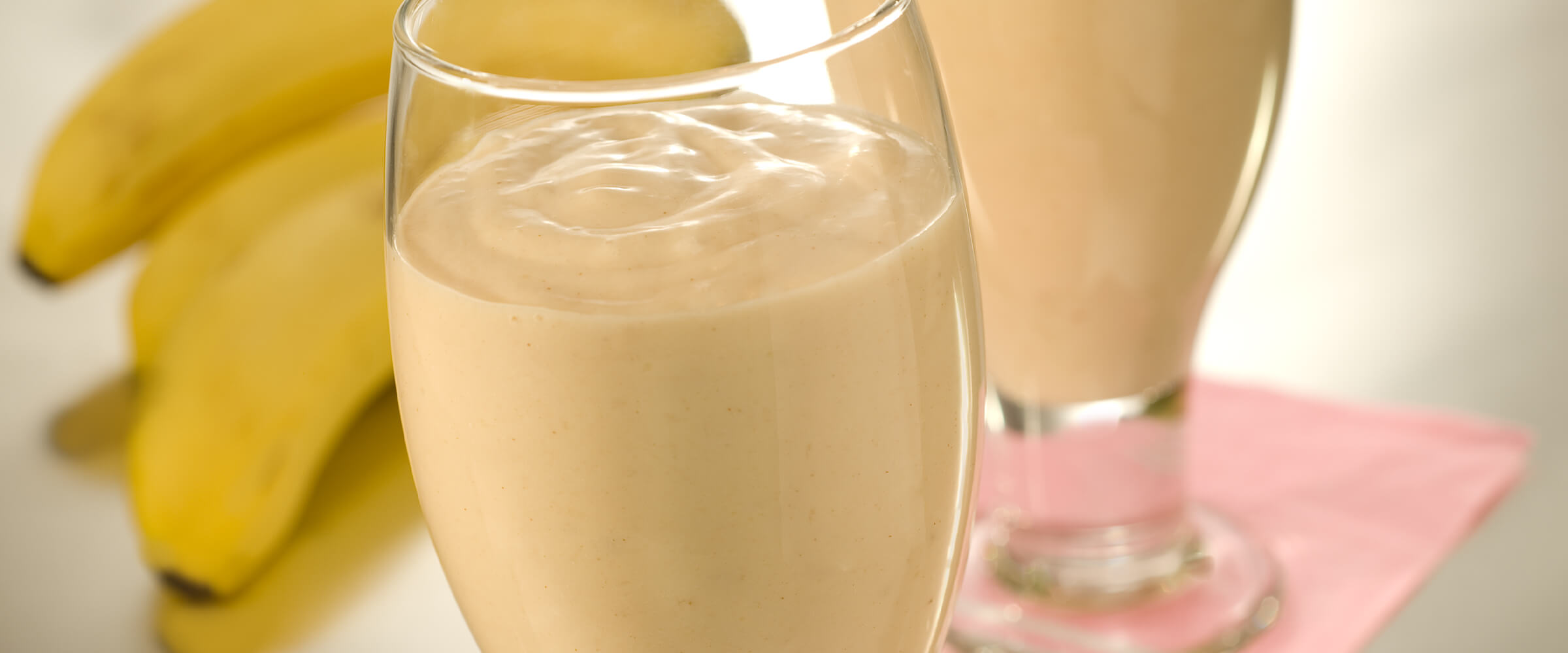 Two glasses of banana breakfast drink with a bunch of bananas behind the two glasses.
