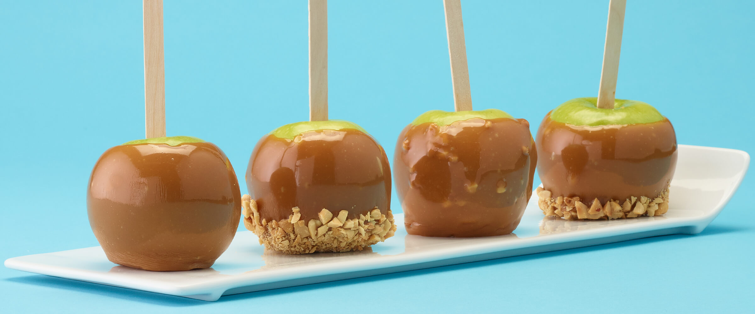 Four caramel peanut butter apples on a teal background sitting on white serving plate.
