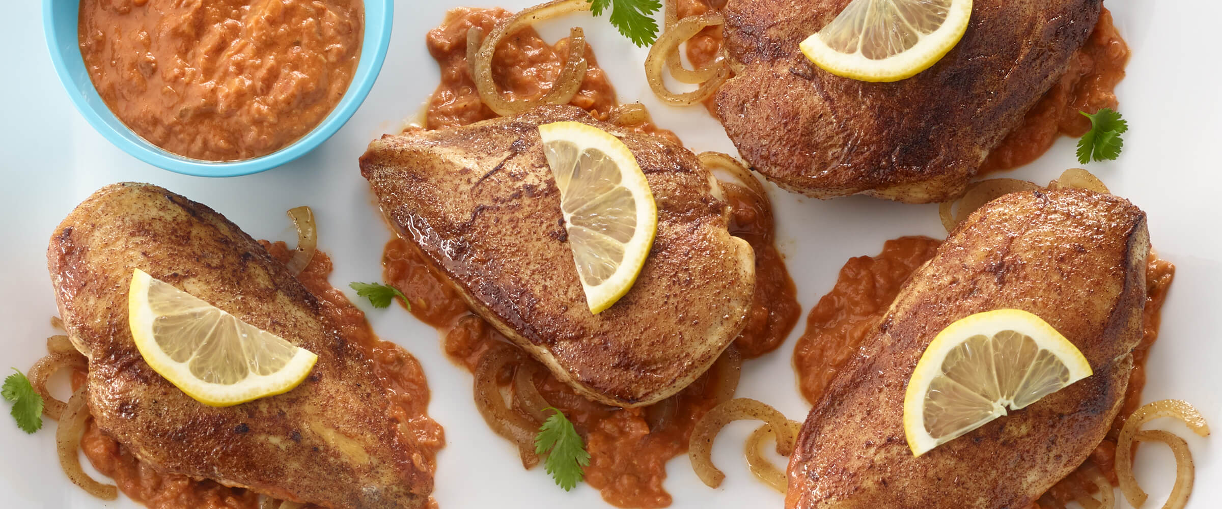 Four chicken breasts in spicy peanut sauce on a large white platter. A small teal dish of salsa is on the platter next to the chicken.