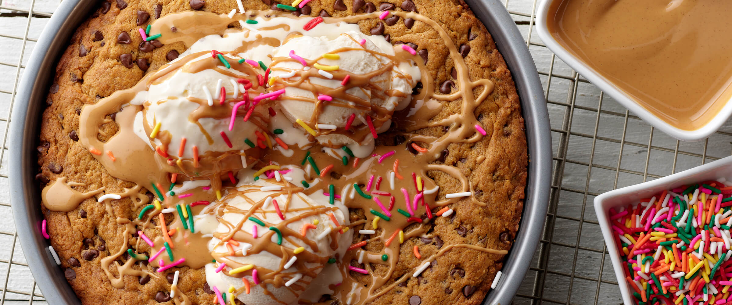 A chocolate chip pumpkin cookie skillet in a large round silver pan with three scoops of ice cream topped with melted peanut butter and multi-colored sprinkles. The pan is on a wire rack with two small bowls of sprinkles and peanut butter sauce.