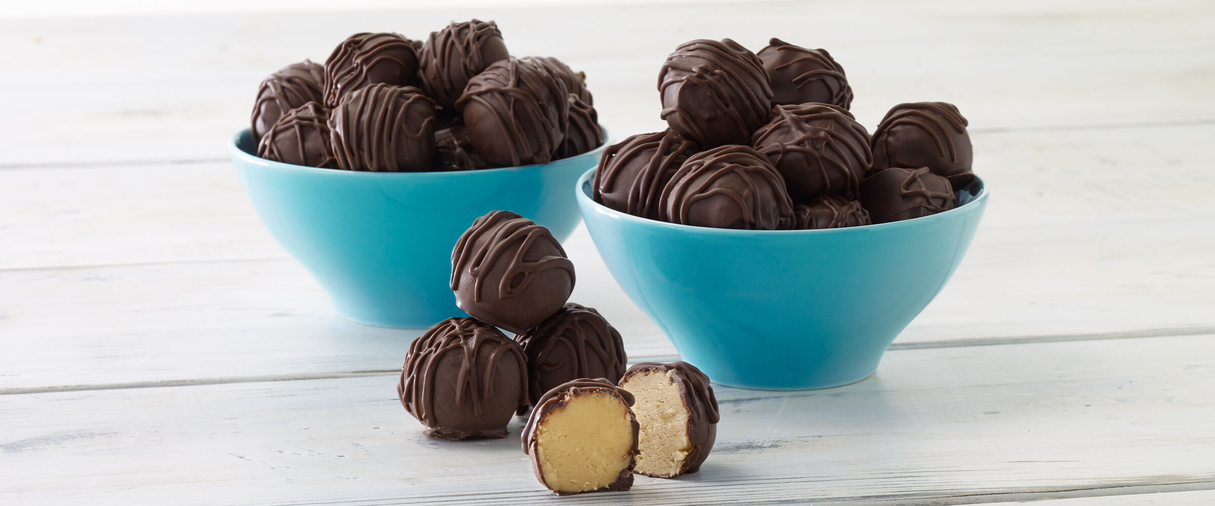 Chocolate covered peanut butter balls in two small, teal bowls sitting on a table with one peanut butter ball in front cut in half.