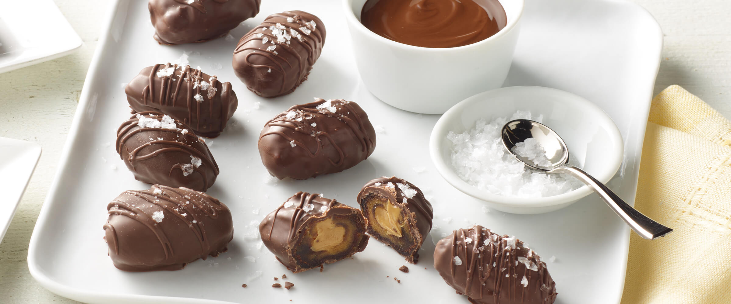 Eight chocolate covered peanut butter dates on a square white plate. Two small bowls are on the plate, one has sea salt and the other has melted chocolate.