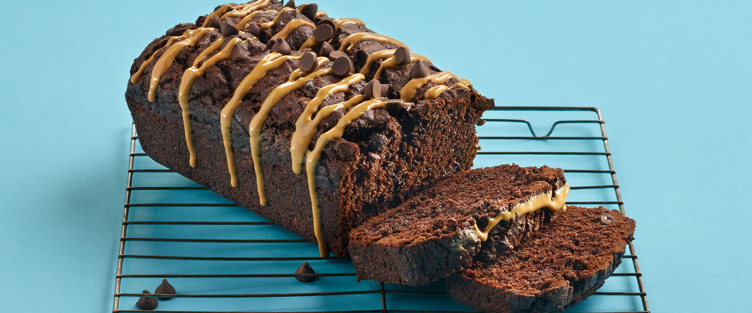 A loaf of chocolate peanut butter banana bread with two slices cut off. The loaf is sitting on a rack on a teal background.