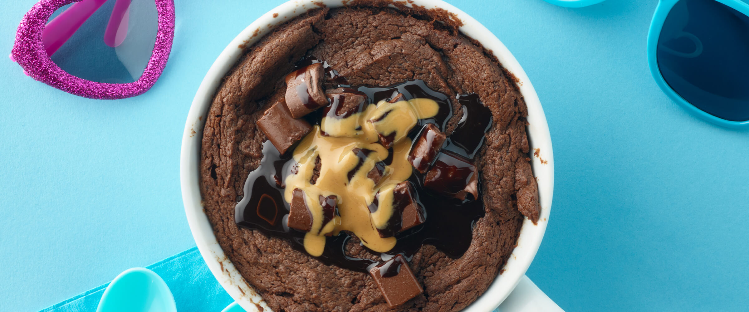 A chocolate peanut butter mug cake in a white mug with pink heart sunglasses and teal sunglasses sitting around the mug.