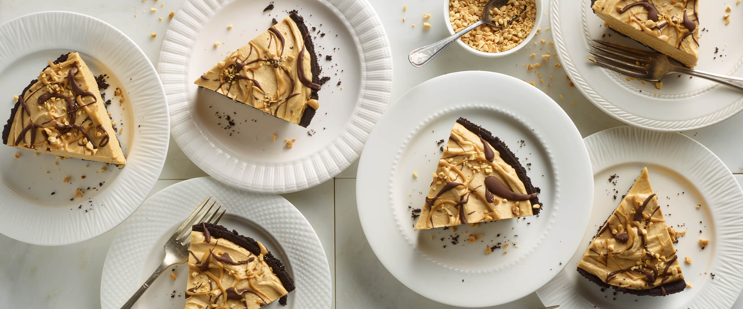 Six slices of chocolate peanut butter pie on six small white plates with forks. A small bowl of crushed peanuts sitting on a white counter top.