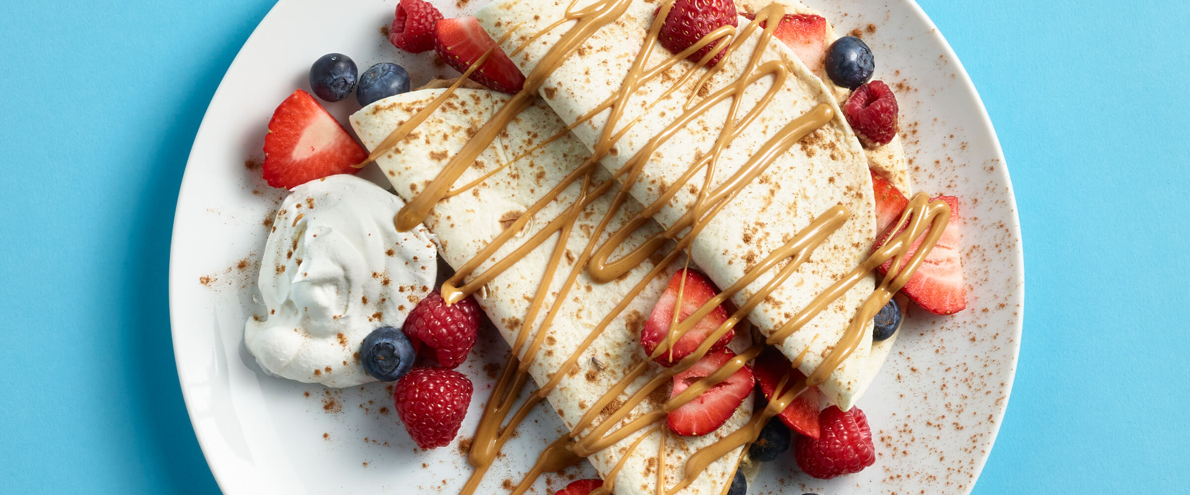 Two fruit tacos on a white plate, with strawberries and blueberries drizzled with peanut butter.