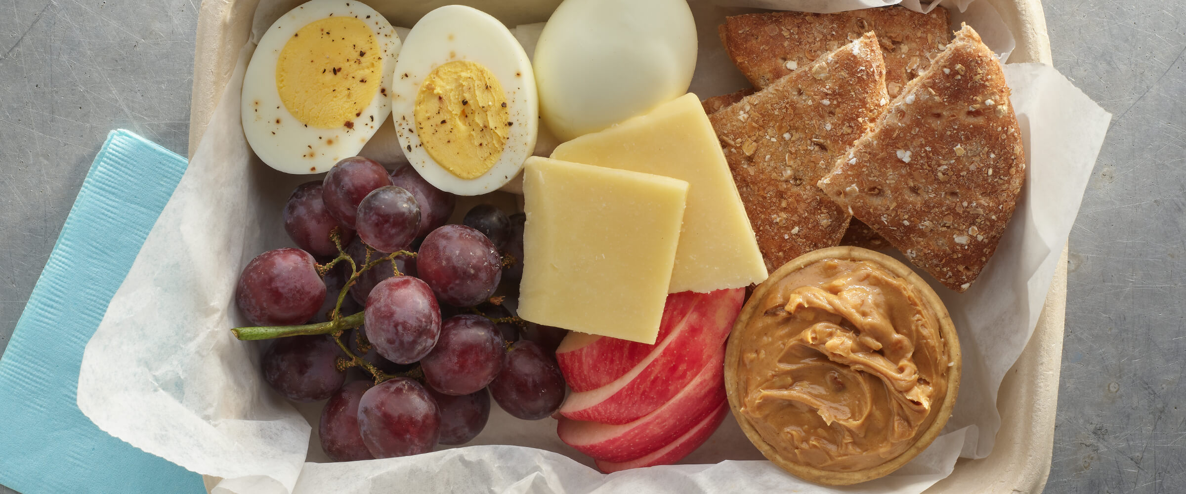 Hard boiled egg, two slices of cheese, red grapes, slices of apple, wheat rounds and a small contained of peanut butter in a rectangular to-go container.