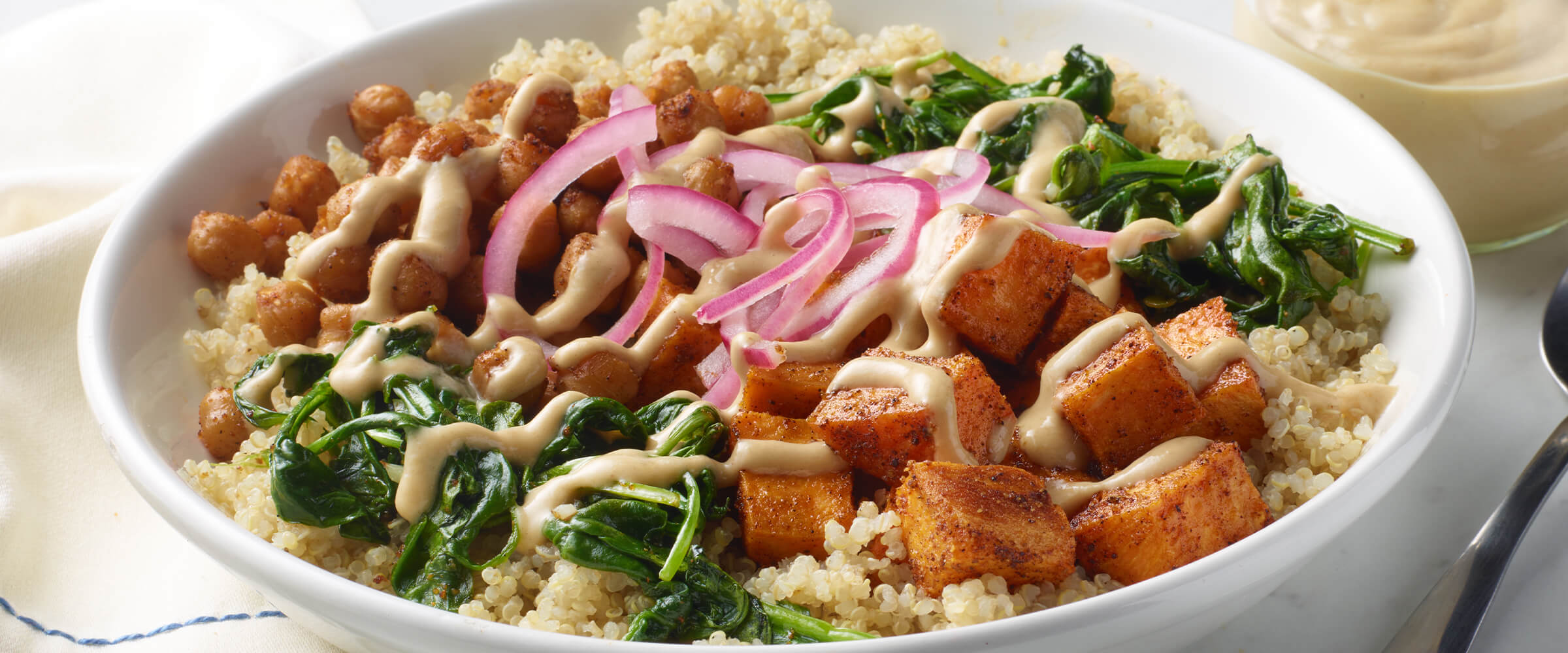 A grain bowl in a white dish with sweet potatoes, quinoa, dressing, spinach and onion.