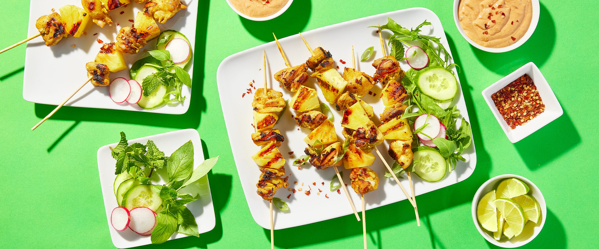 Grilled chicken satay and pineapples on a large white plate with cucumbers, radish and greens. Bowls of lime wedges and red pepepr flakes, along with peanut sauce are around the plate of satay.