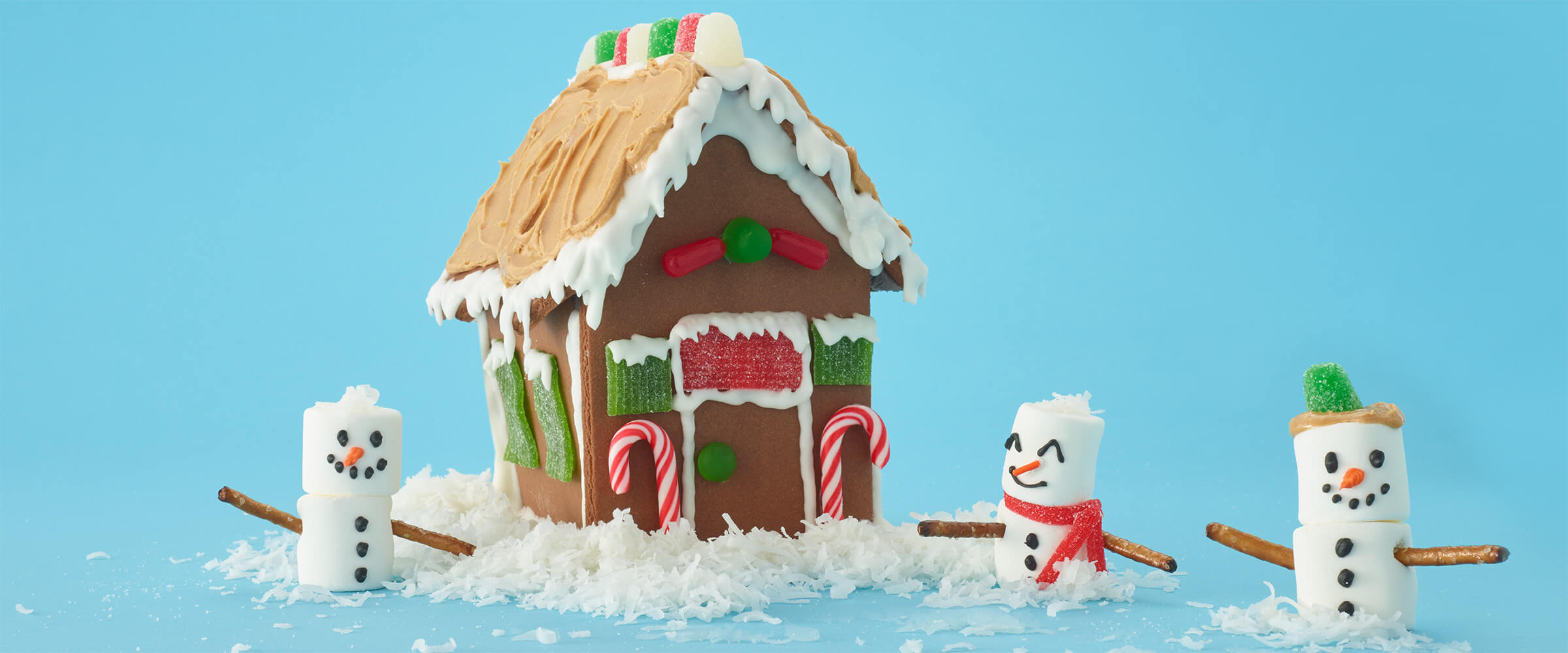 marshmallow men standing in front of a ginger bread house with a peanut butter roof