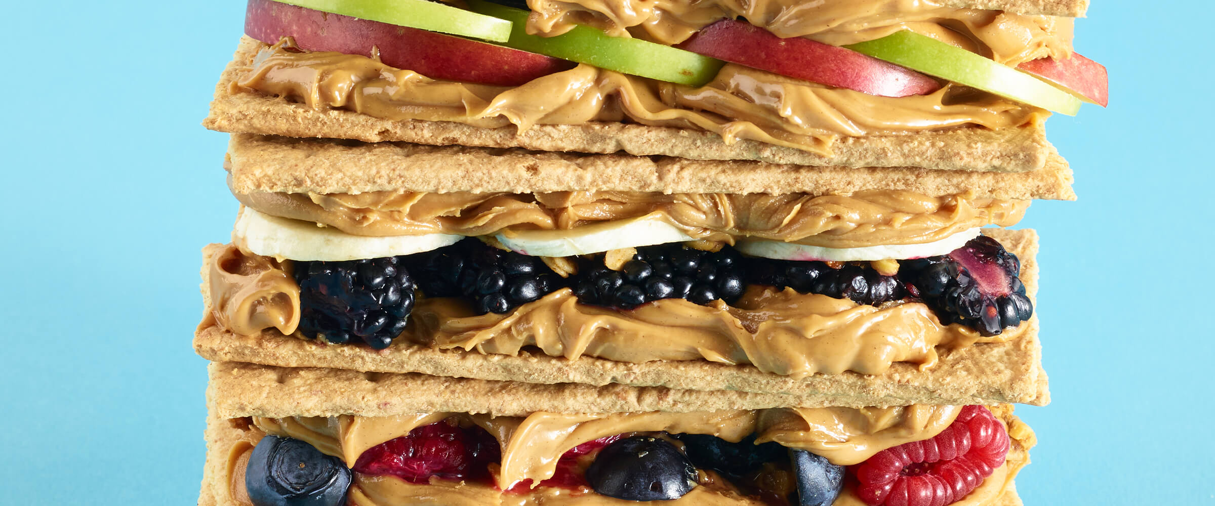 Three PB Sandwich Snacks including peanut butter, apples, grapes and blackberries stacked on top of each other with a teal background.