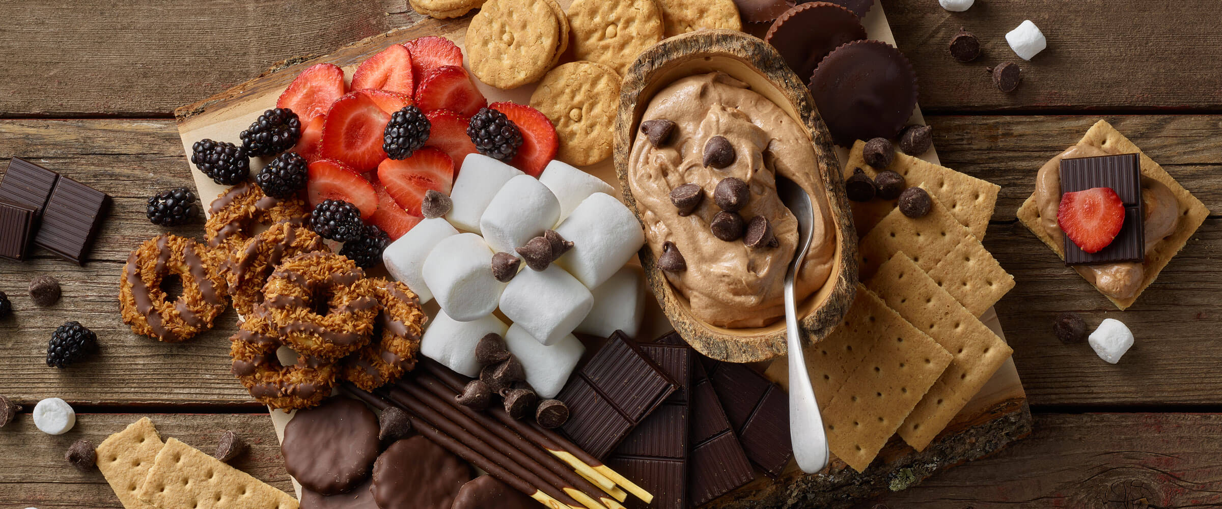 Smores dip, surrounded by chocolate treats, berries, graham crackers and other dippable snacks on a picnic table style surface