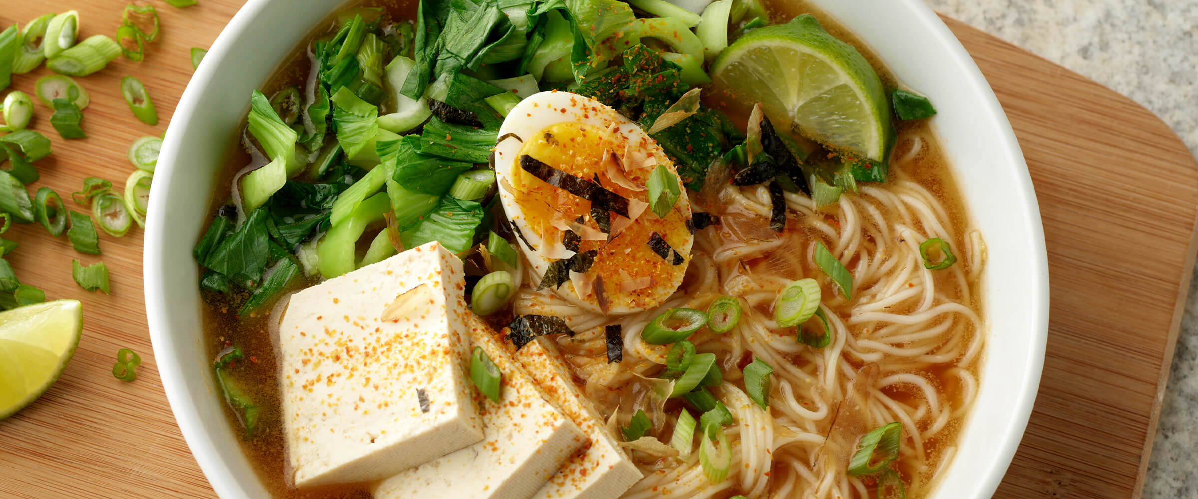 Peanut-Miso Ramen Noodle Soup in a large white bowl. Tofu, a soft-boiled egg, wedges of lime, cilantro and green onion garnish the soup. The bowl is sitting on a wooden board with lime wedges and cut green onion.
