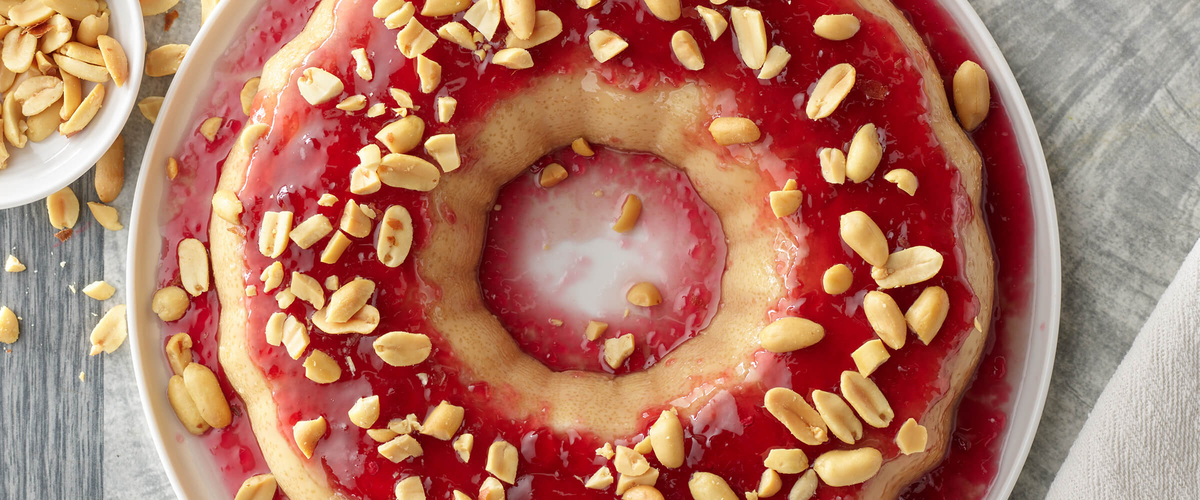 Peanut butter and jelly flan on a large white circular plate. A gray napkin is tucked underneath the plate and a small bowl of peanuts is next to the flan.