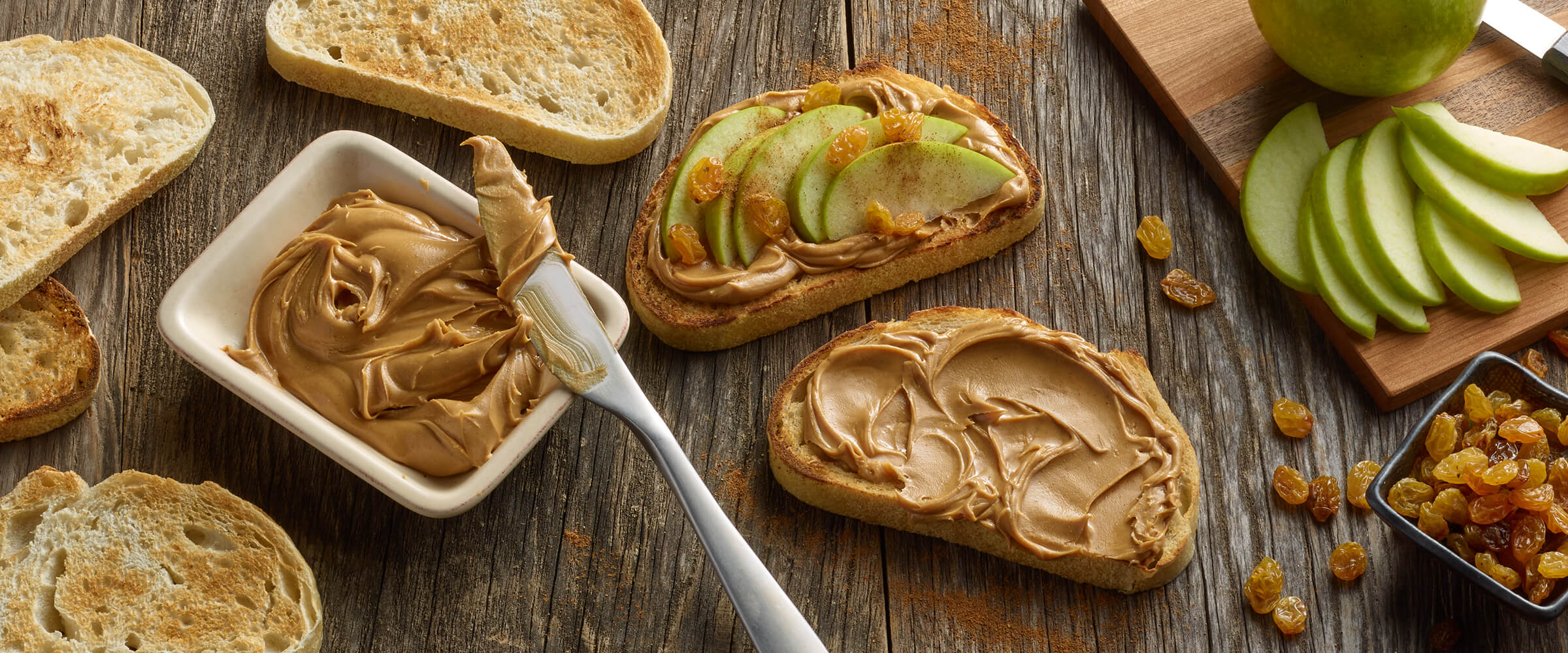 Peanut butter, apple and raisin sandwich sitting on a wodden table. Three slices of bread and a small dish of peanut butter with a knife are to the left pf the sandwich. Slices of green apple on a wooden board are to the left, with a square dish of golden raisins.