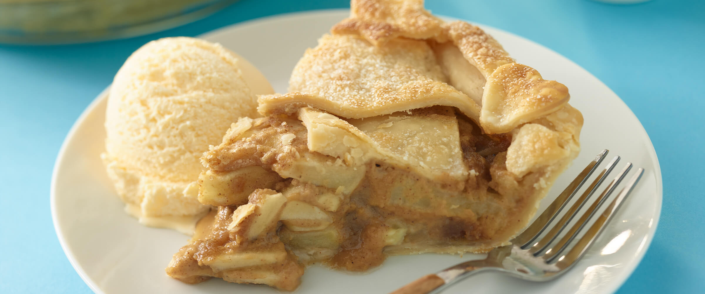 Peanut butter apple pie with a scoop of vanilla ice cream and a fork all on a small white plate sitting on a teal background.