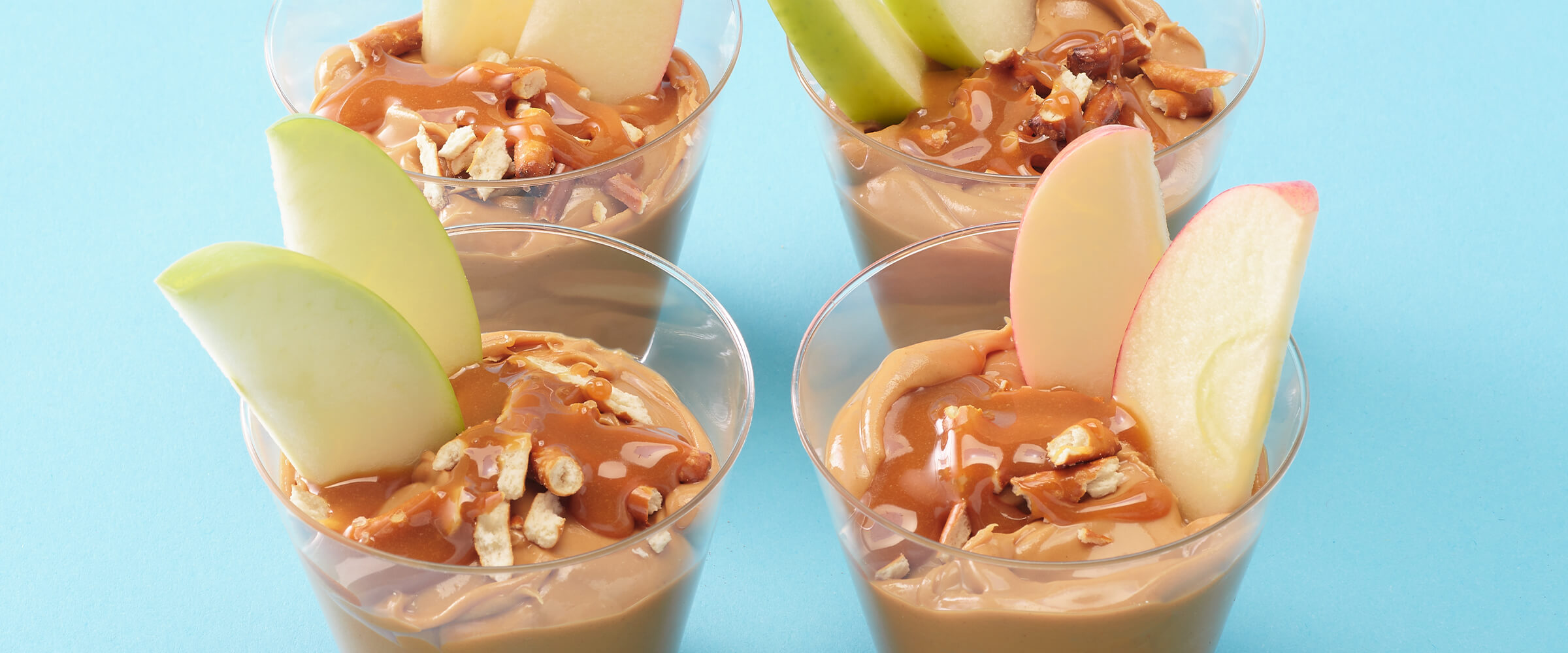 Four peanut butter apple snack cups. The peanut butter is topped with crushed pretzels and caramel sauce, and has two apple slices in each clear cup.