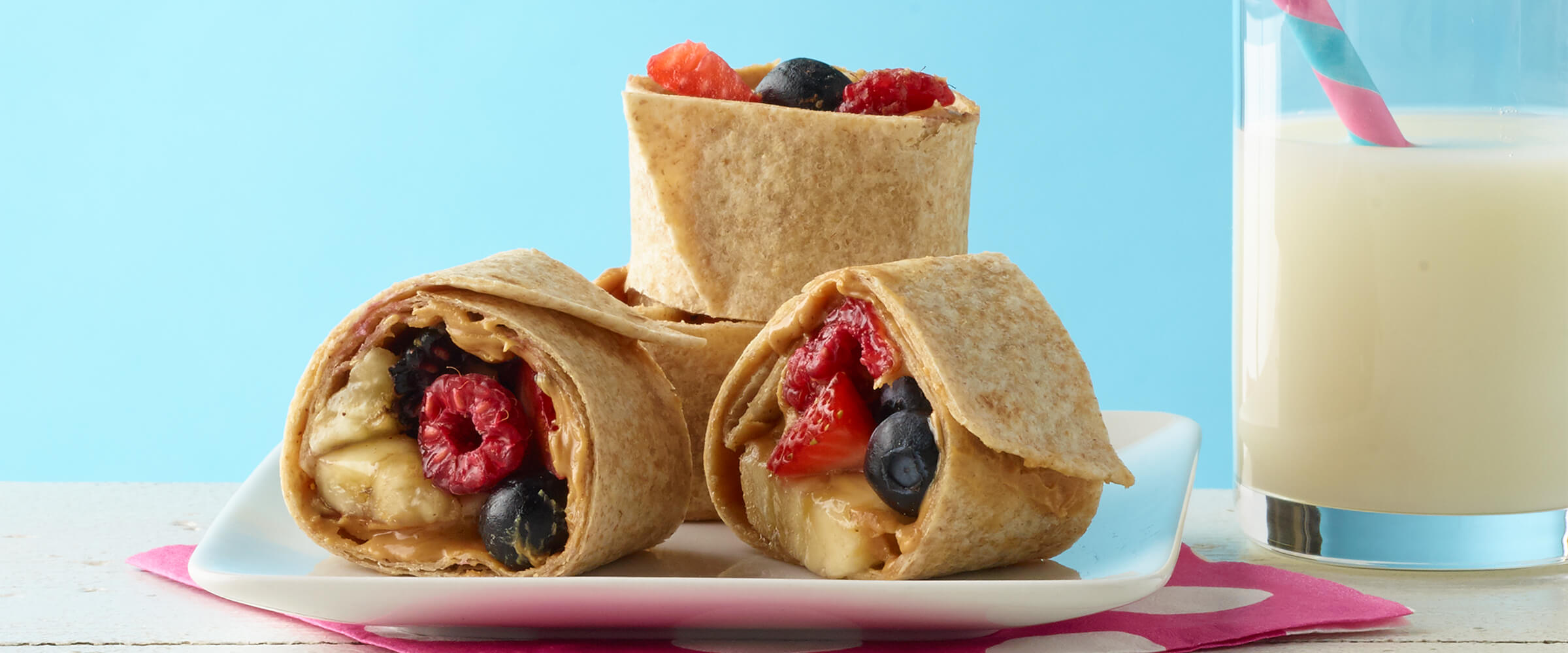Three peanut butter banana berry spiral sitting on a white square plate. A glass of milk with a pink and blue spiral straw is next to the plate.