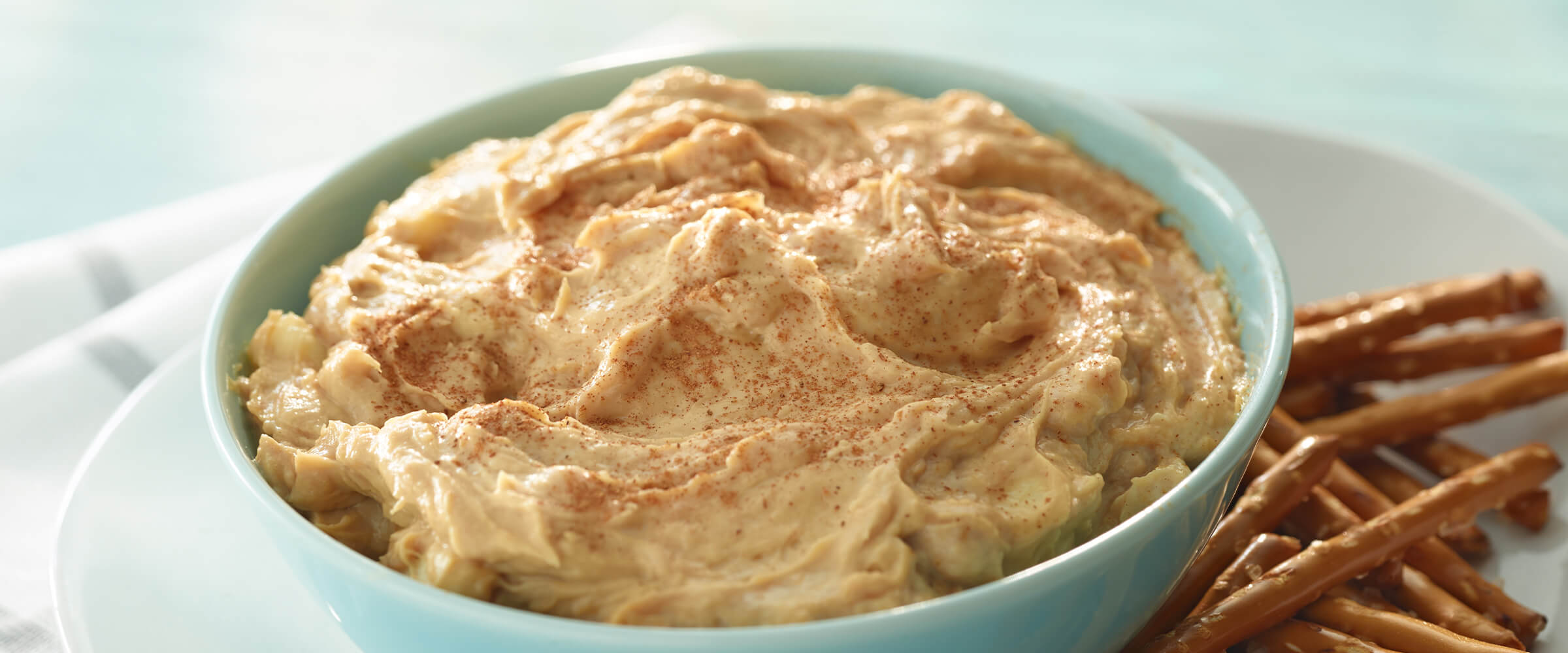 Peanut butter banana dip in a teal bowl sitting on a plate with pretzel sticks.