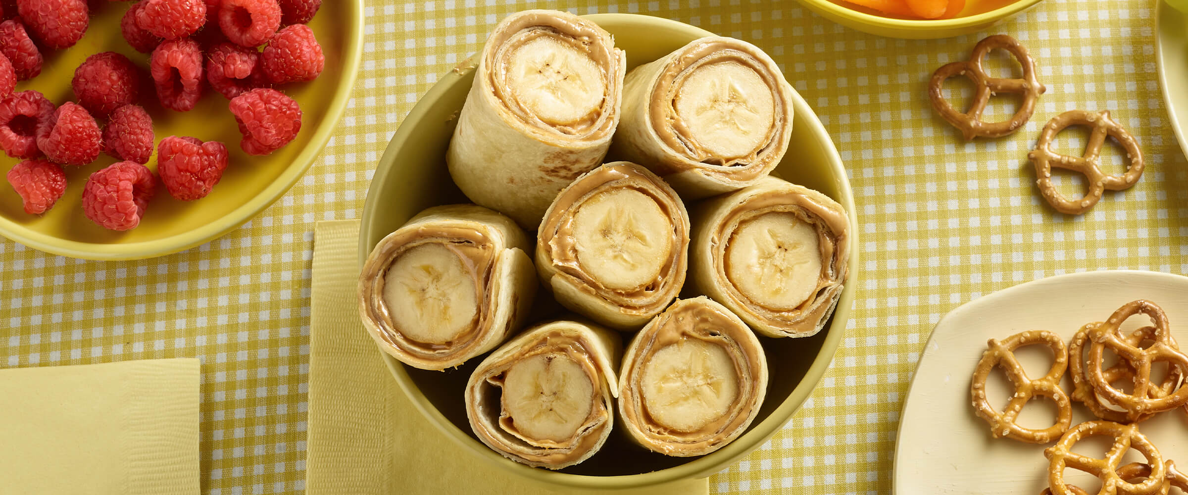 Peanut butter banana roll-ups in a yellow bowl with a plate of pretzels and a bowl of rasperries. All sitting on a table with a yellow and white plaid tablecloth.