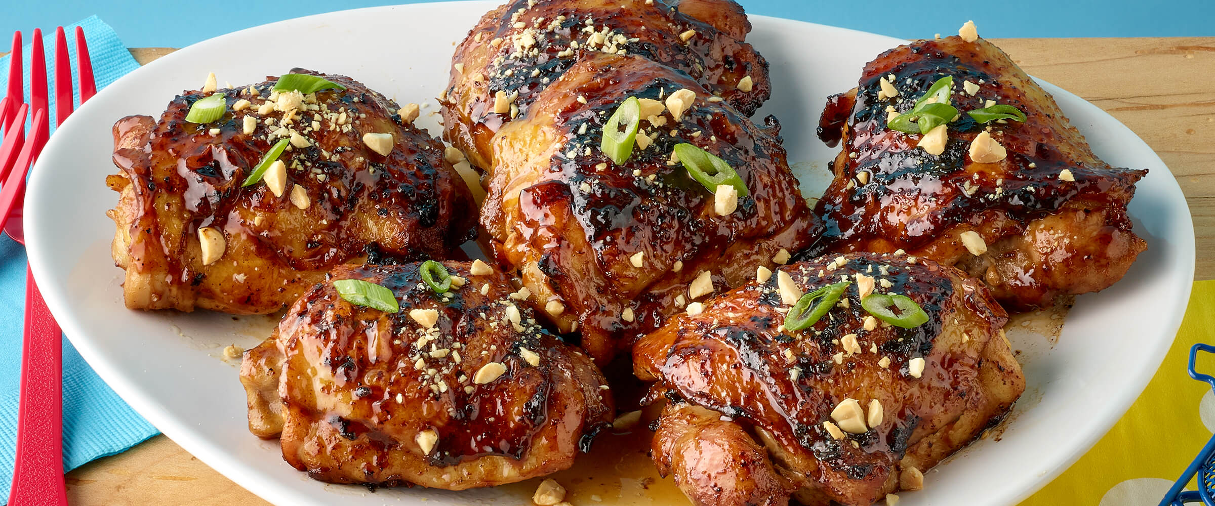 Six pieces of grilled peanut butter chicken on a large white plate. Red forks on a teal napkin tucked under the white plate.