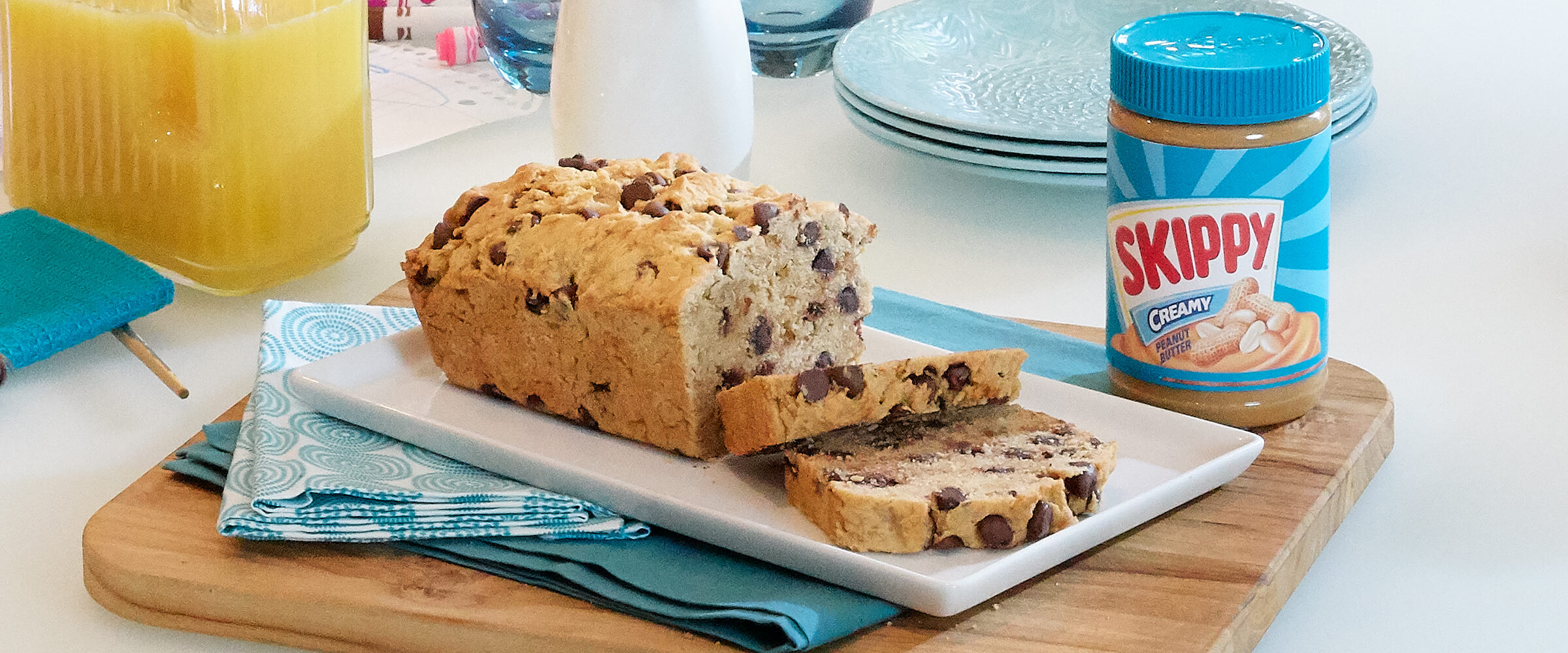 A loaf of peanut butter chocolate chip bread with two slices cut off the end, sitting on a white serving plate with a teal jar of SKIPPY® peanut butter next to the plate