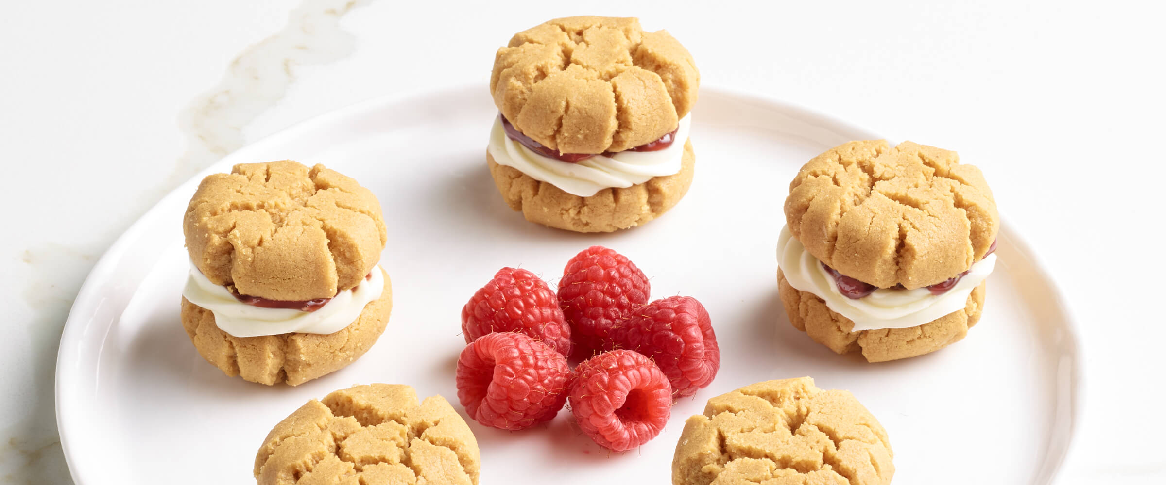 Five peanut butter cookie sandwiches on a white plate with five raspberries in a flower shape in between all of the cookie sandwiches.