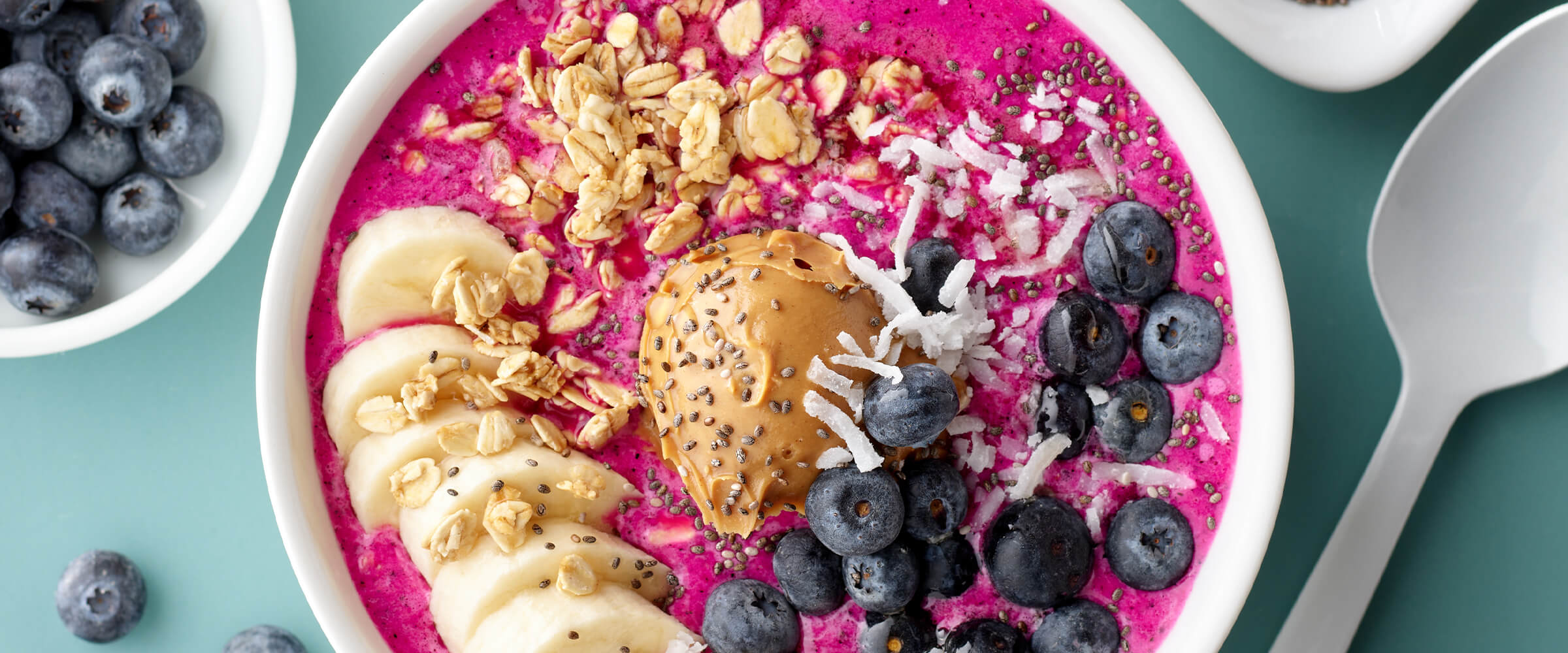 Peanut butter dragon fruit bowl topped with peanut butter, blueberries, coconut, granola and bananas. The bowl is on a teal counter with a smal bowl of blueberries and a large spoon nearby.