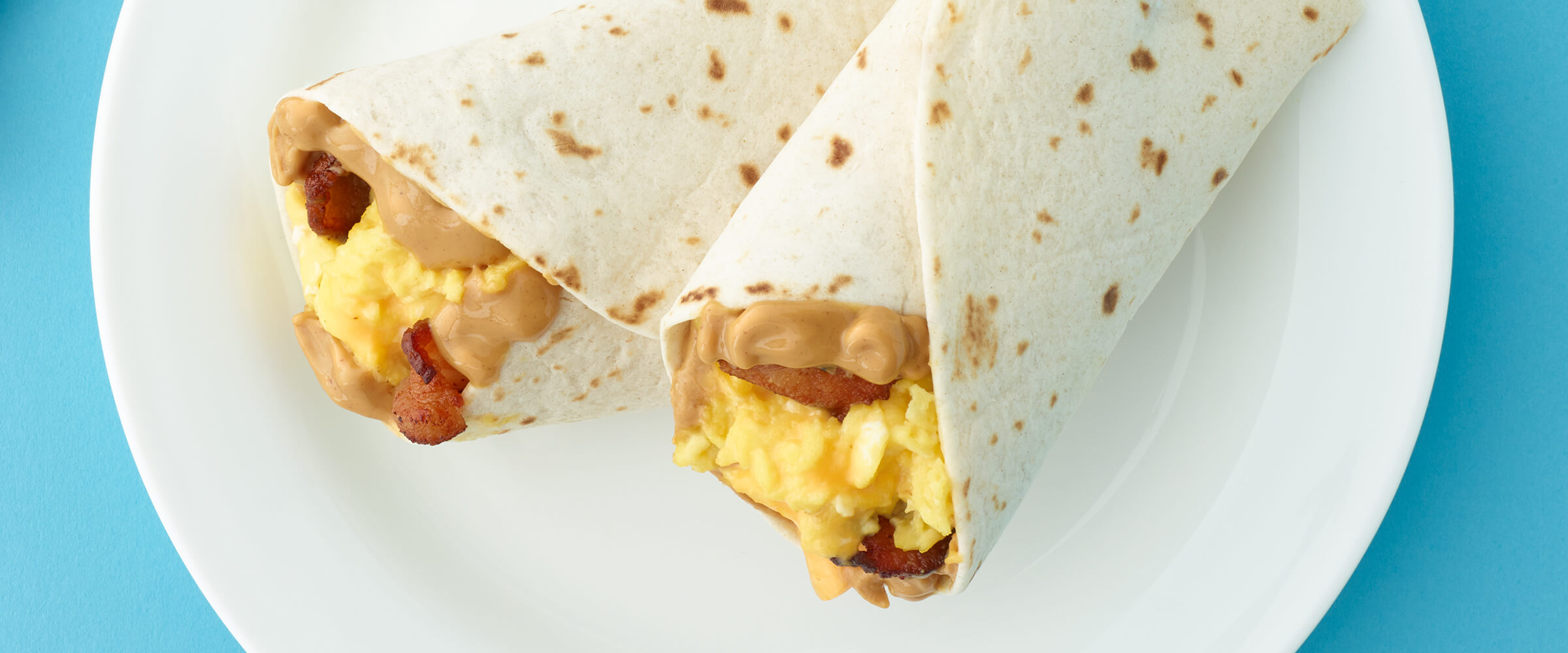 Two peanut butter, bacon, egg and cheese wraps on a white plate.