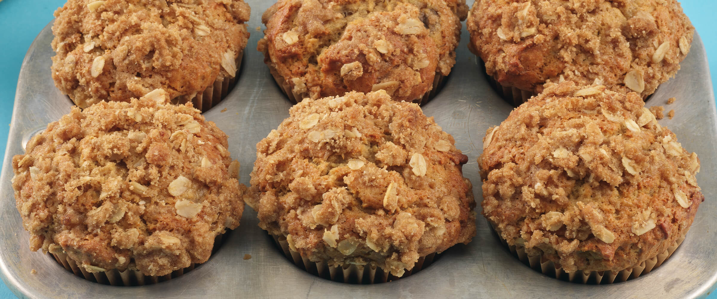 Six peanut butter energy muffins in a muffin tin on a teal background.