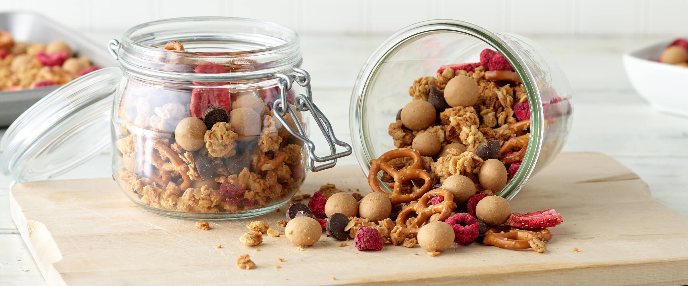 Two jars of peanut butter granola trail mix on a wood board, one is tipped over with the trail mix spilling out