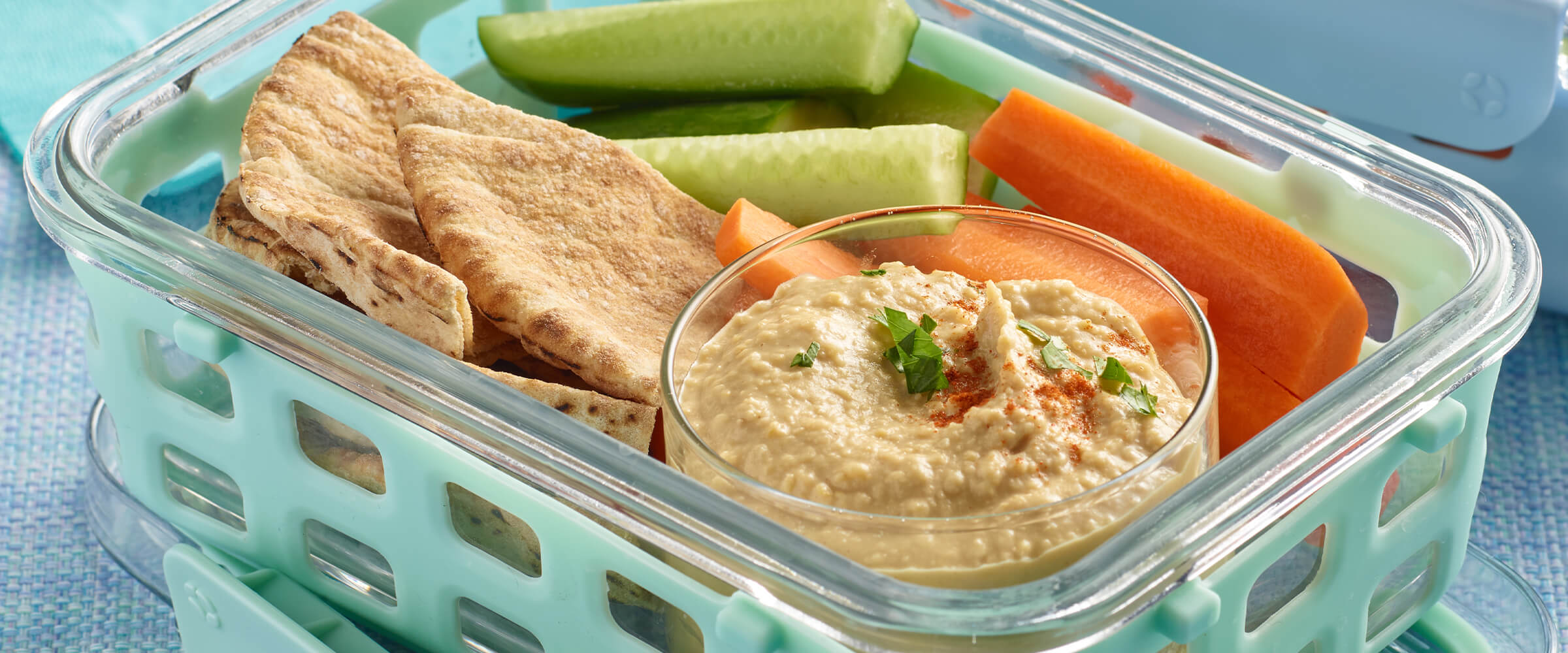 Peanut butter hummus in a small dish. The dish of hummus is inside a teal glas container with cut up pita bread, sliced carrot and celery sticks.