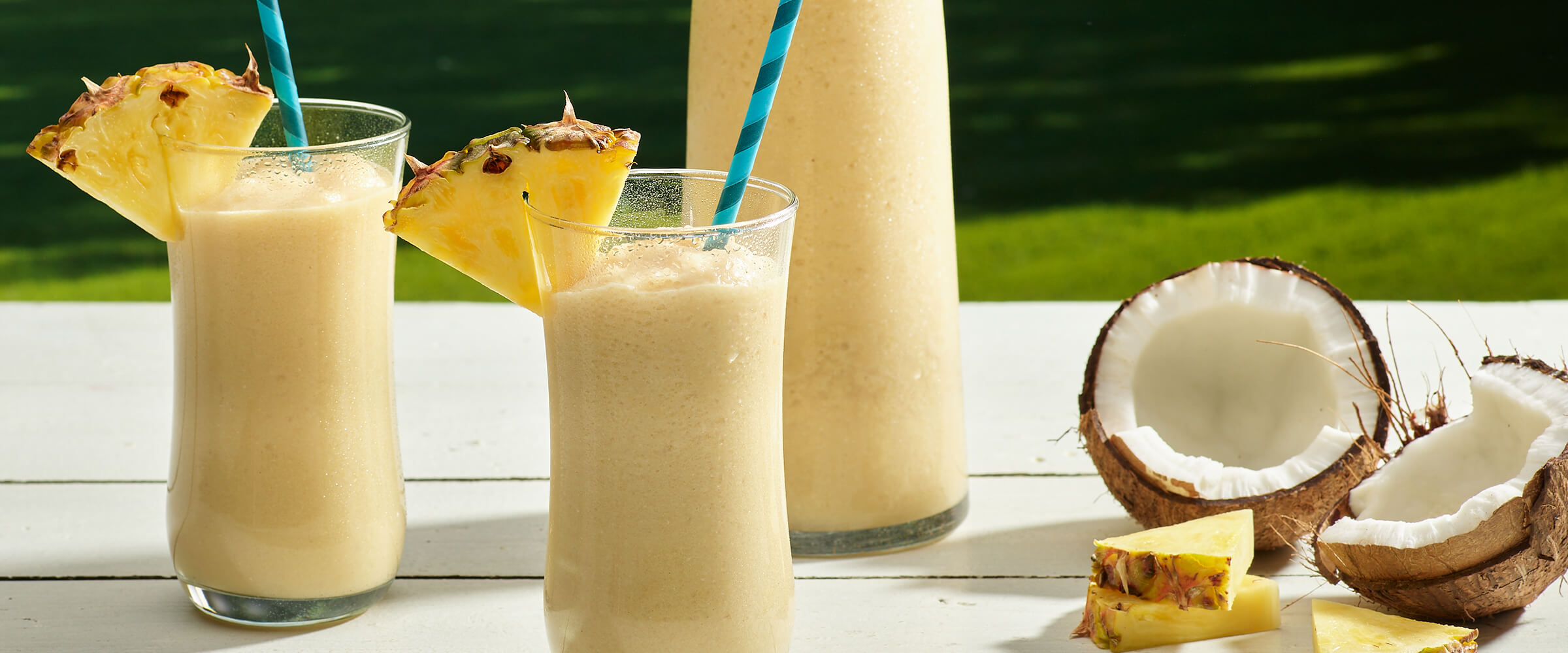 Peanut butter pina colada in two glasses with wedges of pineapple on the rim. A broken coconut and pineapple wedges are next to the glasses, along with a pitcher of pina colada.