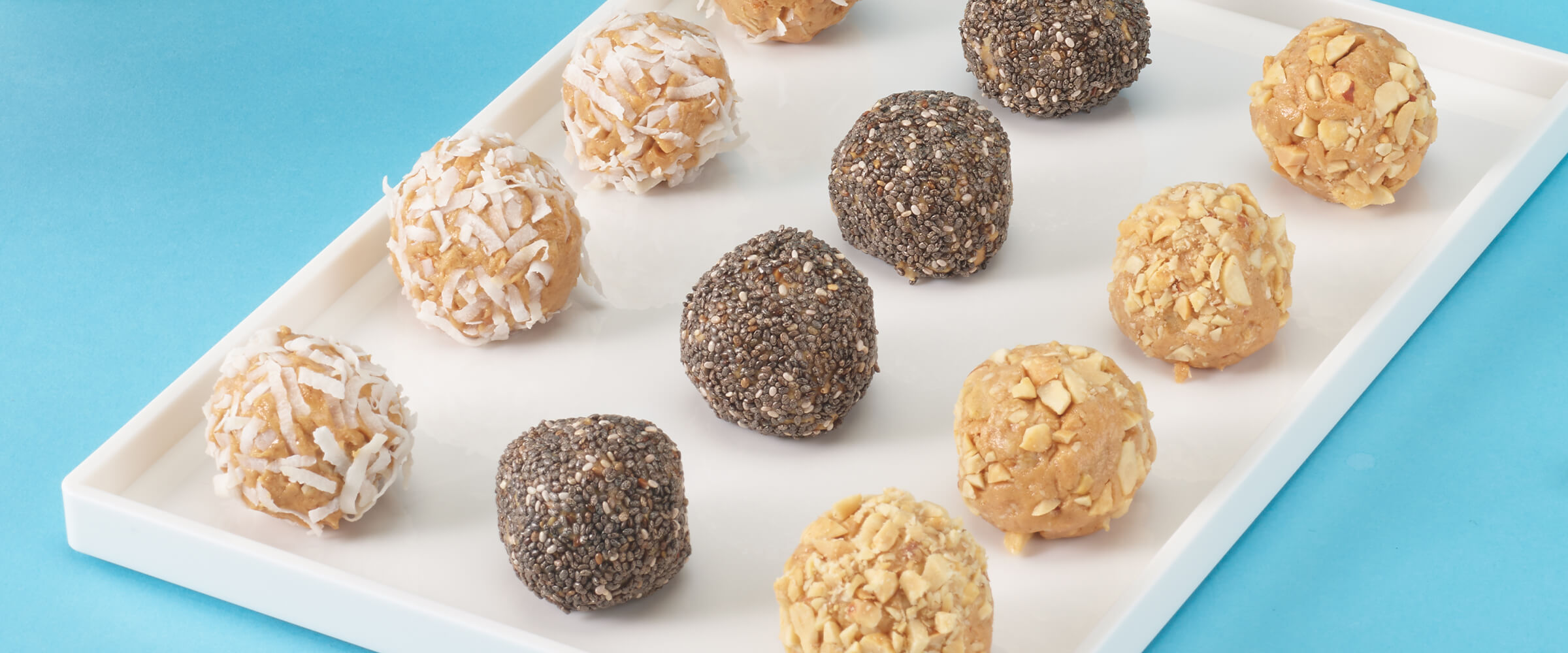 12 peanut butter proteins balls on a rectangular white serving platter. Four of them are covered in coconut, four are covered in chocolate and four are covered in crushed peanuts.