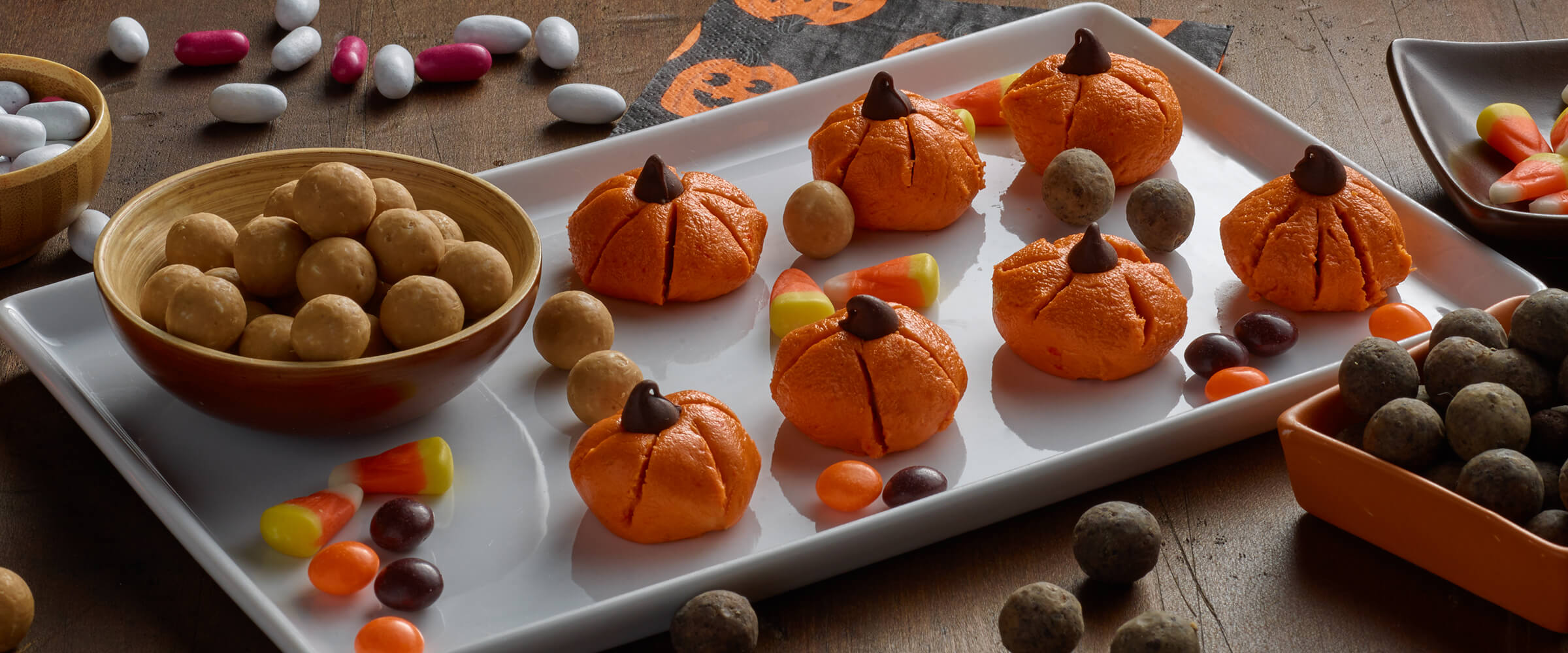 Seven peanut butter pumpkins sitting on a white rectangular serving plate with candy corn and P.B. Bites throughout the plate.