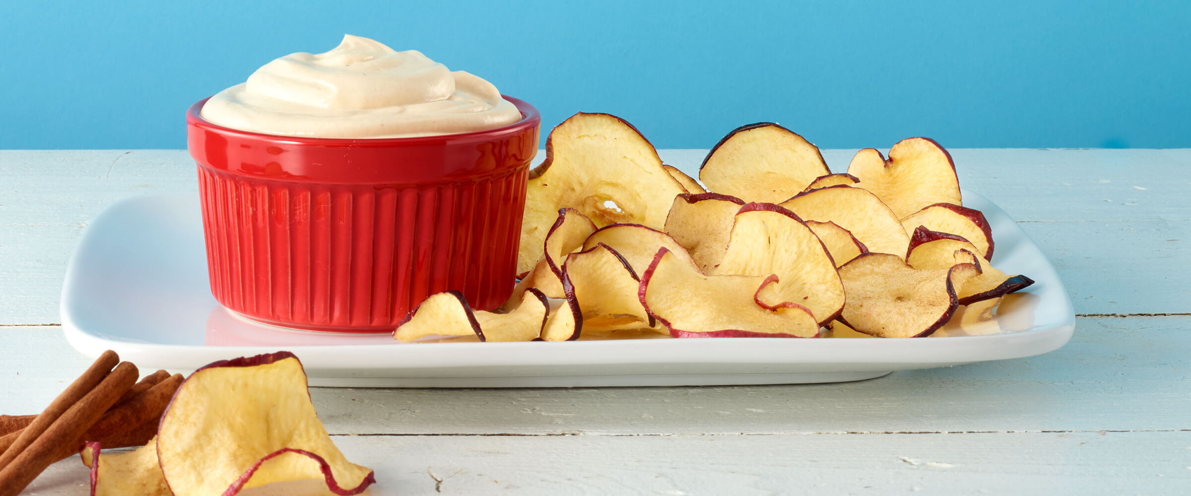 Peanut butter yogurt sauce in a small red dish. The dish is sitting next to apple chips on a rectangular white plate.