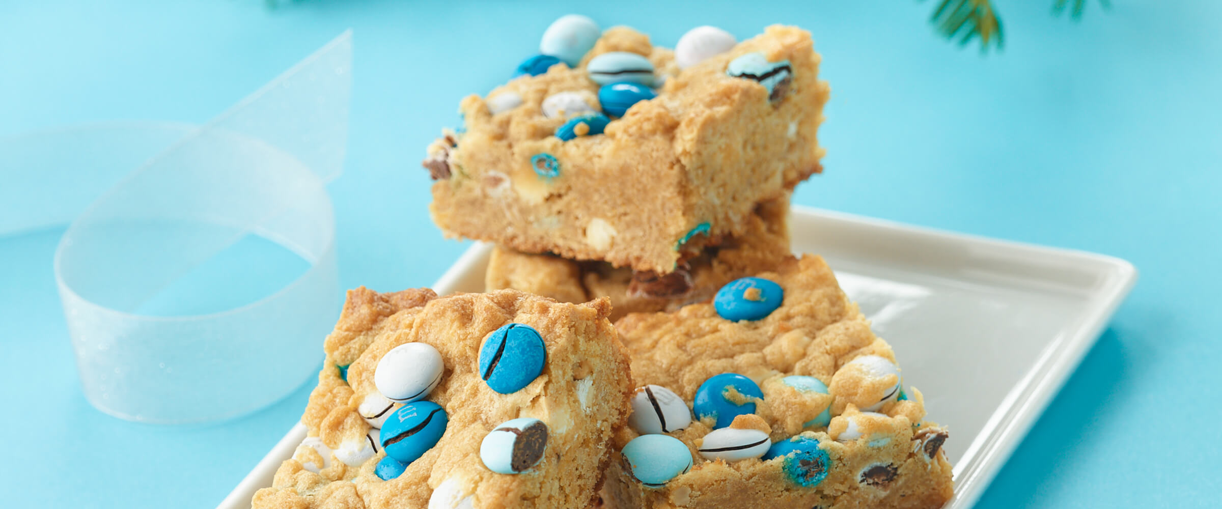 Three peanut butter and candy cookie bars stacked on top of each other on a square white plate with a teal background.