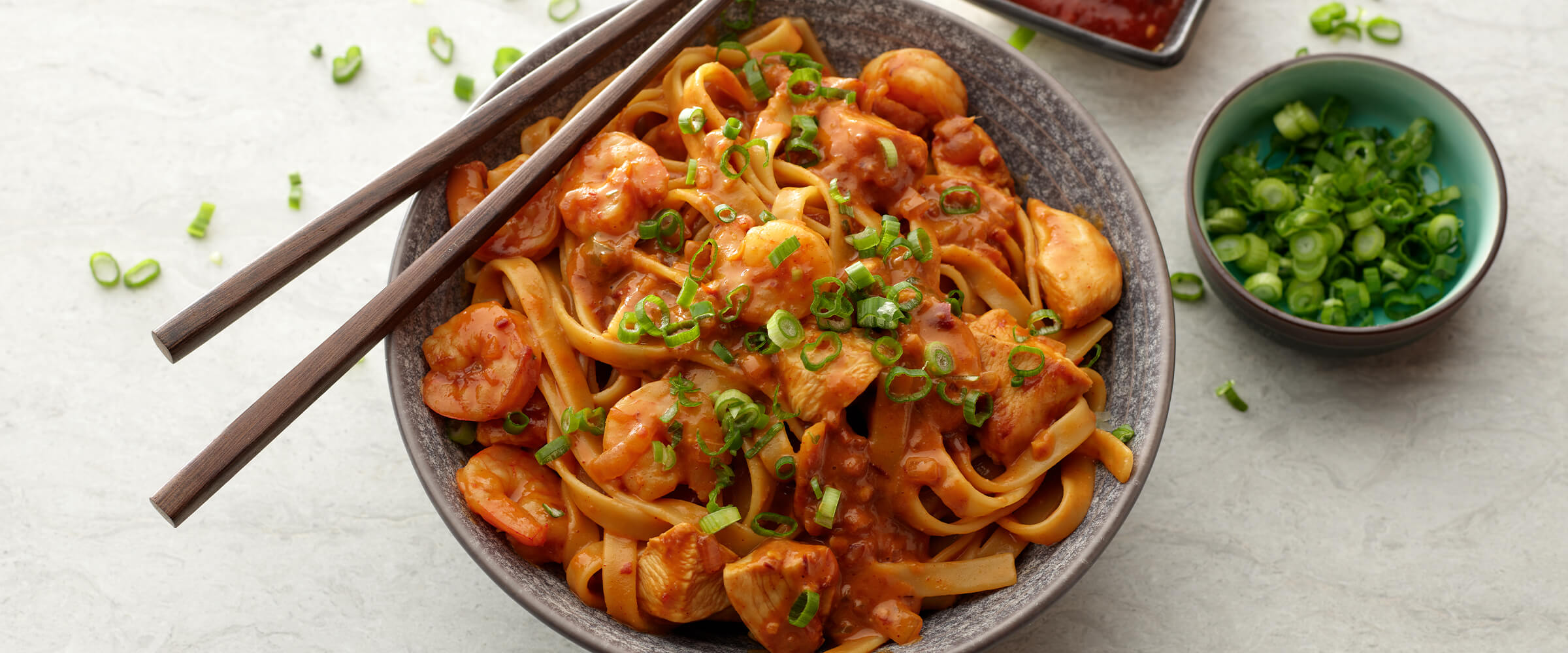 Peanut butter and chili garlic noodles in a large bowl with chopsticks resting on top. A small bowl of green onion and chili oil sits next to the bowl of noodles.