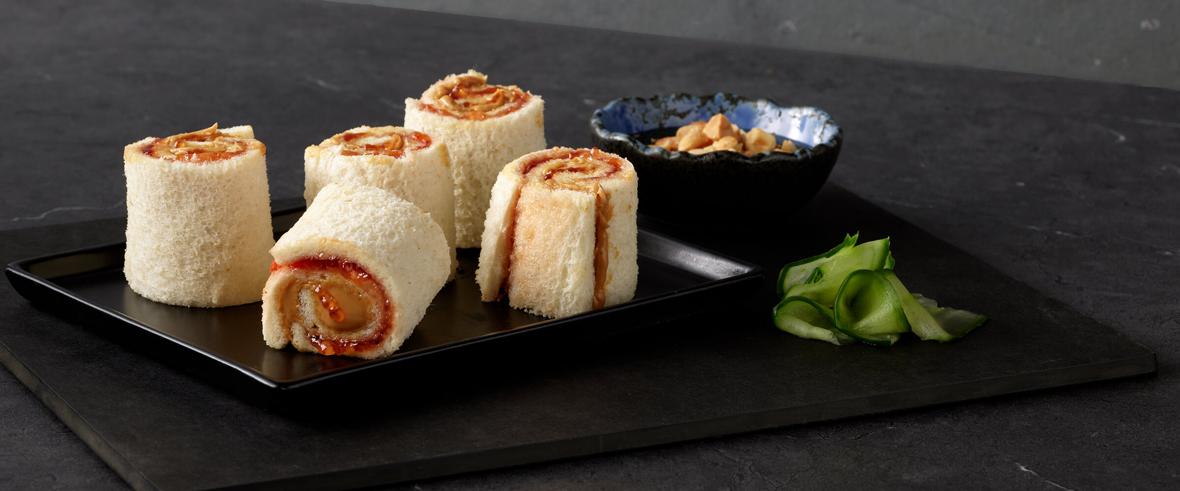 Five peanut butter and jelly suhi rolls on a black plate with a small bowl of peanuts and a sprig of mint