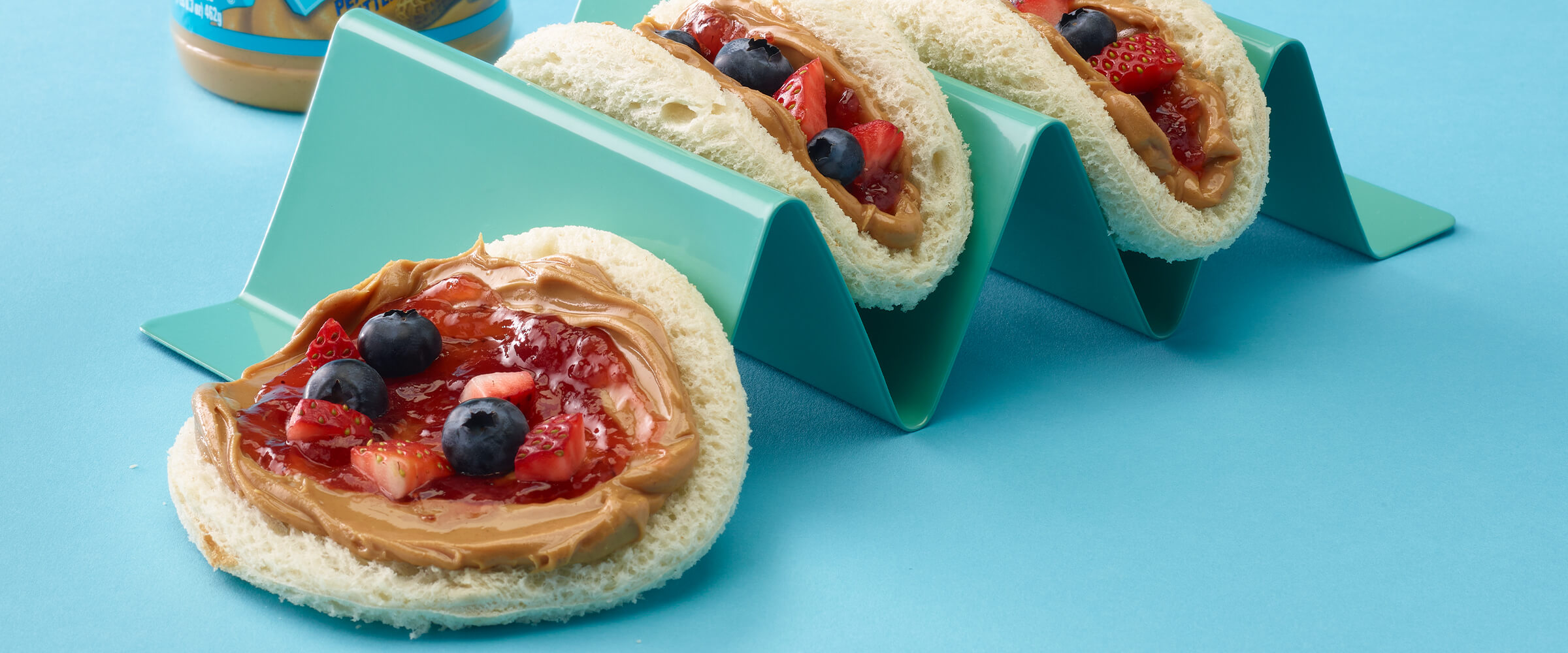 Two peanut butter and jelly tacos in a teal taco holder. One taco is off to the side of the holder on a teal background.