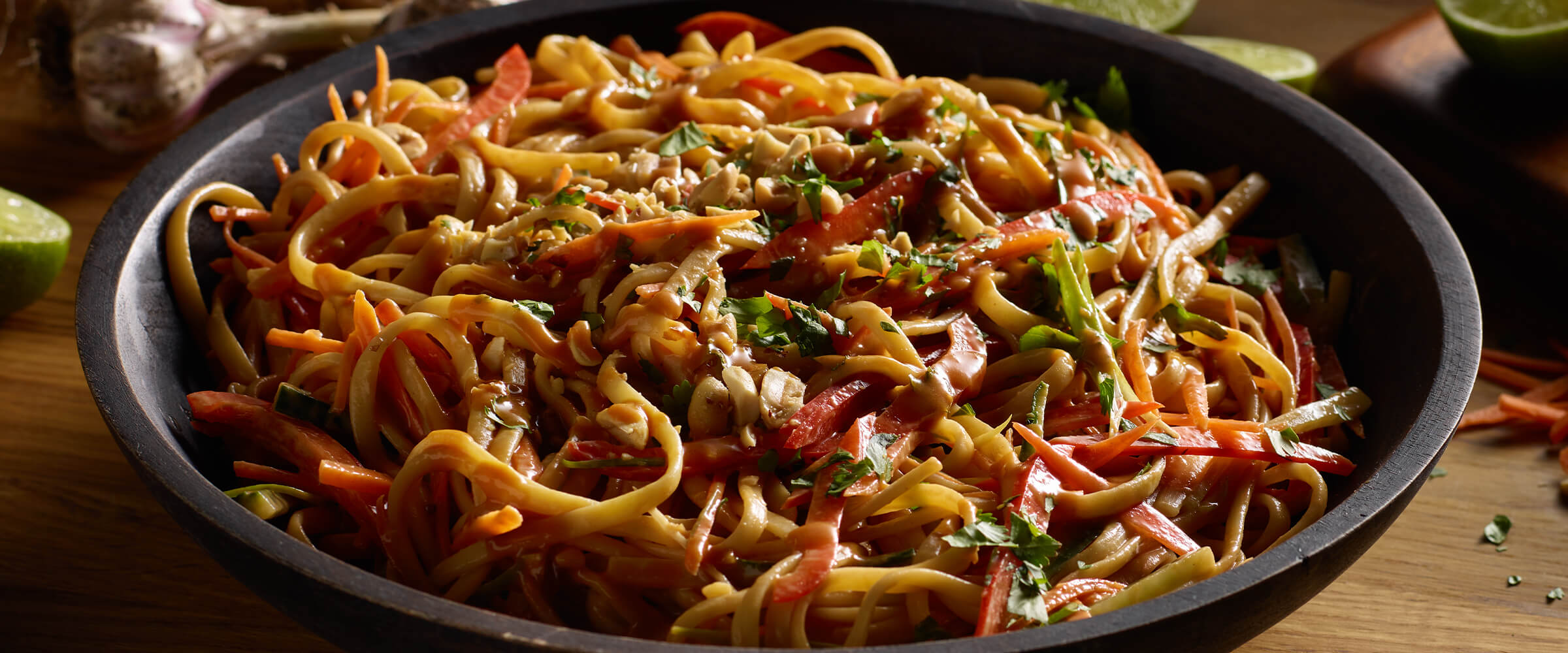 Peanut noodles with sliced carrot, red bell pepper, cilantro and green onion in a large black bowl sitting on a wooden board.