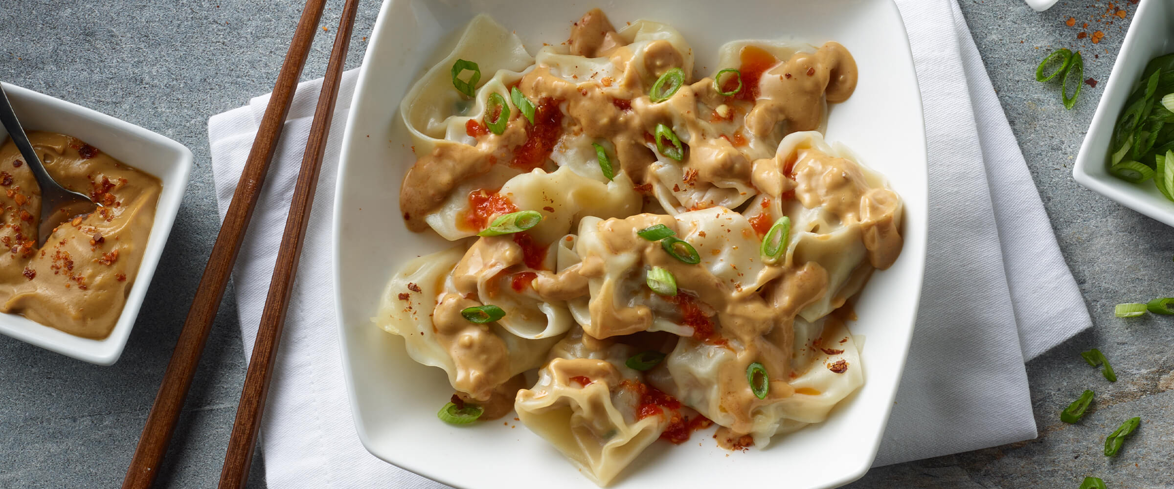 Wontons with spicy peanut sauce in a large white bowl with chopsticks and a bowl of peanut sauce sitting off to the left.