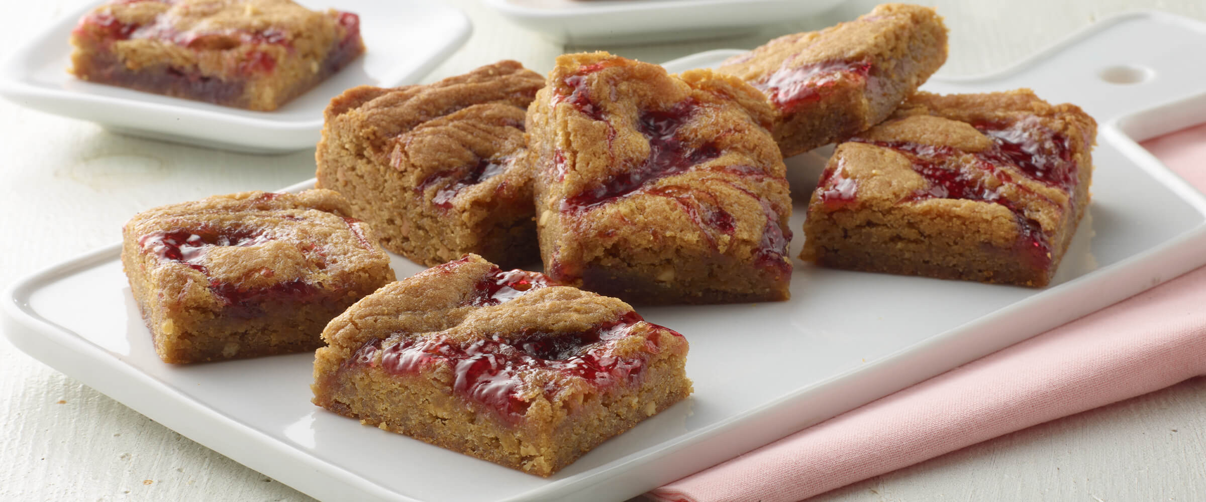 Peanut butter and jelly blondies stacked on a white platter with a pink napkin tucked underneath